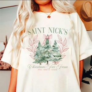 May include: Cream-colored t-shirt featuring the text "SAINT NICK'S Christmas Tree Farm" in green script. The design includes watercolor-style Christmas trees, a pink bow, and floral accents. The shirt is a relaxed fit.