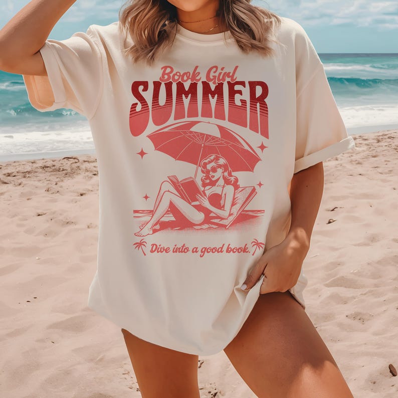 May include: A white t-shirt with a pink graphic of a woman reading a book under an umbrella on the beach. The text on the shirt reads "Book Girl Summer" and "Dive into a good book."