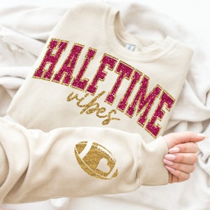 May include: A beige sweatshirt with the text "HALFTIME with you" in pink glitter and a gold glitter football with a heart on the sleeve.