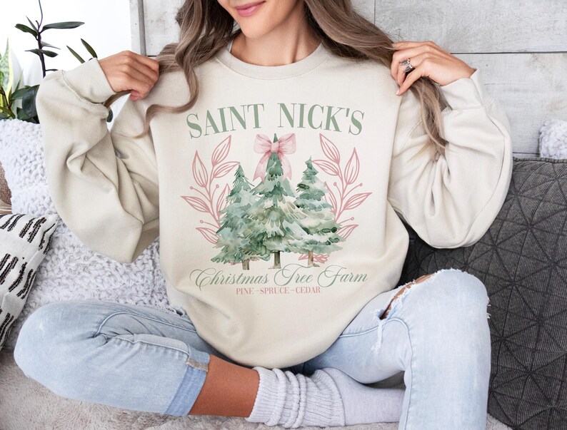 May include: Cream-colored sweatshirt with the text "Saint Nick's Christmas Tree Farm" and a watercolor illustration of three Christmas trees. The design includes a pink bow and floral accents. The sweatshirt is a relaxed fit.
