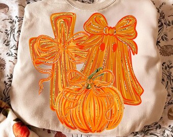 May include: Cream-colored sweatshirt featuring a Halloween-themed design. The design includes an orange pumpkin and two orange ghosts with bows, all outlined with gold glitter. A festive and seasonal apparel item.