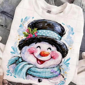 May include: A white crewneck sweatshirt featuring a graphic illustration of a smiling snowman wearing a black top hat with a light blue polka dot band, green holly leaves, and red berries, paired with a light blue scarf and pink cheeks.