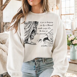 May include: White sweatshirt with a black and white illustration of two figures and the text "if you're hanging on by a thread make sure it's the hem of his garment. Matthew 9-20-27"