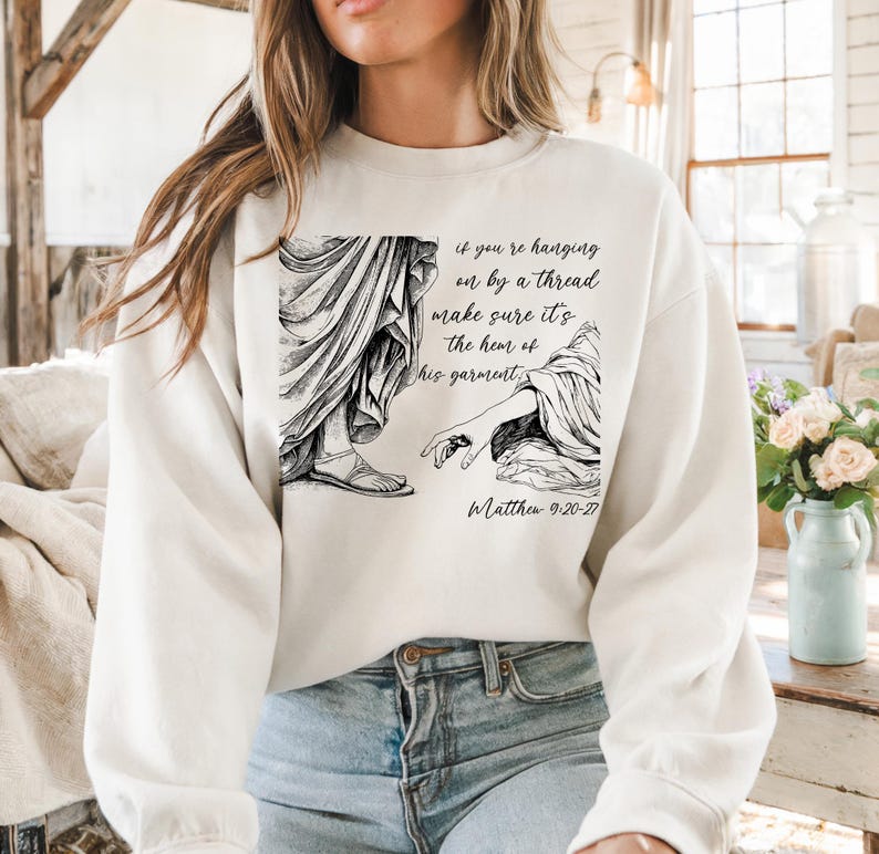 May include: White sweatshirt with a black and white illustration of two figures and the text "if you're hanging on by a thread make sure it's the hem of his garment. Matthew 9-20-27"