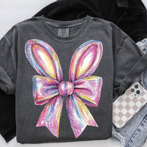 May include: A dark gray t-shirt with a large, colorful, sparkly bow design. The bow is pink, purple, yellow, and blue.