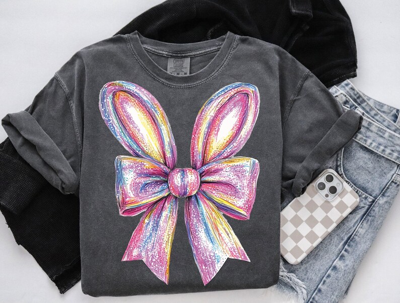May include: A dark gray t-shirt with a large, colorful, sparkly bow design. The bow is pink, purple, yellow, and blue.