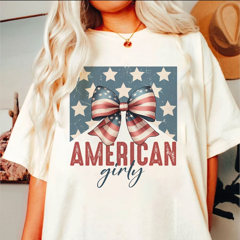 May include: White t-shirt with a red, white, and blue bow with stars in the background. The text "AMERICAN girly" is printed below the bow.
