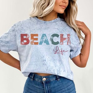 May include: A light blue t-shirt with a distressed look featuring the text "BEACH life" in colorful letters with palm trees. The letters are in shades of red, orange, yellow, green, and blue.