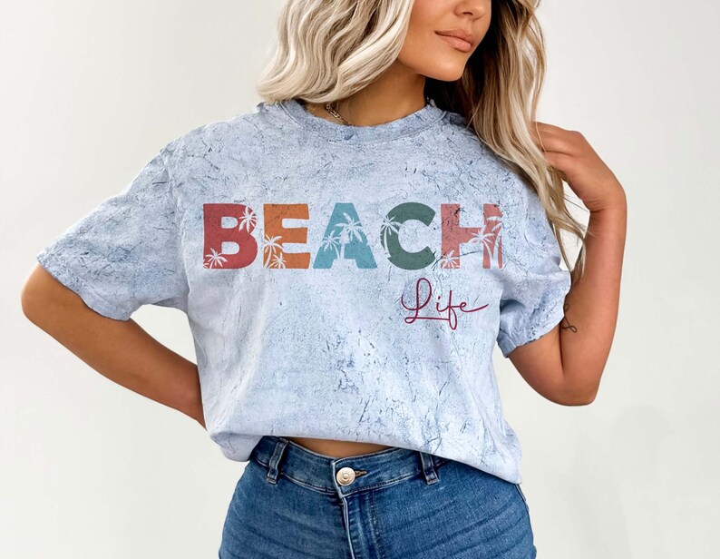 May include: A light blue t-shirt with a distressed look featuring the text "BEACH life" in colorful letters with palm trees. The letters are in shades of red, orange, yellow, green, and blue.