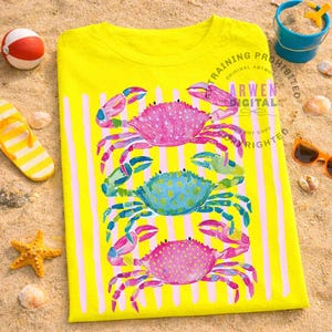 Puede incluir: Camiseta amarilla con rayas verticales rosas y blancas. Presenta tres coloridos cangrejos en rosa, azul y verde. El texto "ARWEN DIGITAL" es visible. Accesorios de playa rodean la camiseta.