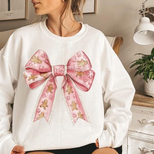 Puede incluir: Sudadera blanca con un gran lazo rosa con hombres de jengibre y copos de nieve. El lazo es el punto focal, con un dise&ntilde;o detallado de acuarela. La sudadera es una prenda c&oacute;moda de manga larga, perfecta para la temporada navide&ntilde;a.