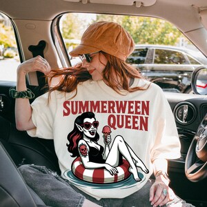 May include: Cream-colored t-shirt with the text "SUMMERWEEN QUEEN" in red. The graphic features a vampire woman in a swimsuit, heart-shaped sunglasses, and a tattoo, holding an ice cream cone while floating on a red and white striped inner tube.