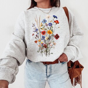 May include: A gray sweatshirt with a floral print featuring a bouquet of colorful wildflowers, including red, yellow, blue, and purple flowers. The sweatshirt has long sleeves and a crew neck.