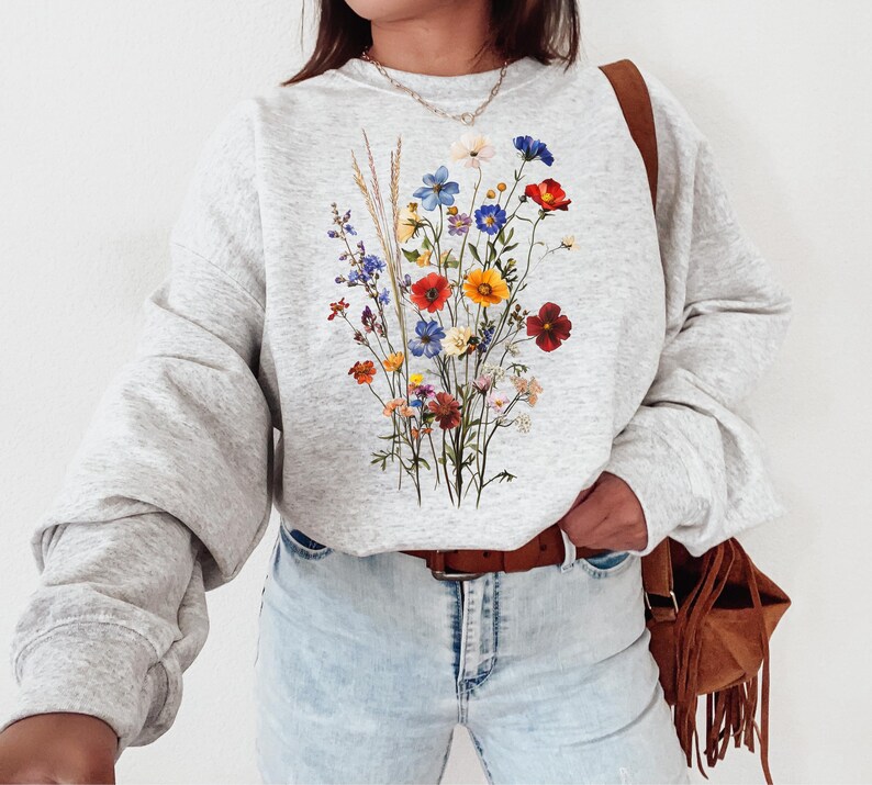 May include: A gray sweatshirt with a floral print featuring a bouquet of colorful wildflowers, including red, yellow, blue, and purple flowers. The sweatshirt has long sleeves and a crew neck.