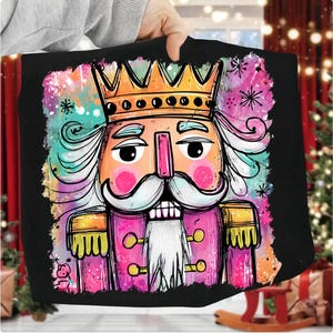 May include: Black tote bag featuring a colorful illustration of a nutcracker soldier with a gold crown, pink jacket, and white beard. The design includes splashes of teal, pink, and orange, with snowflake accents, perfect for holiday gifts.