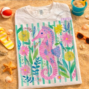 May include: White t-shirt featuring a pink seahorse design with floral accents and vertical green stripes. The seahorse is surrounded by colorful flowers and foliage. The shirt is displayed on a sandy surface with beach-themed accessories.