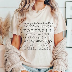 May include: A beige t-shirt with black text listing fall-themed phrases like "cozy blankets" and "football." The shirt is paired with a light-colored cardigan. The overall aesthetic suggests a cozy, autumn-themed style.