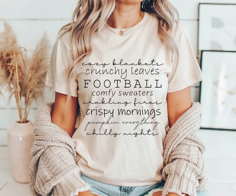 May include: A beige t-shirt with black text listing fall-themed phrases like "cozy blankets" and "football." The shirt is paired with a light-colored cardigan. The overall aesthetic suggests a cozy, autumn-themed style.