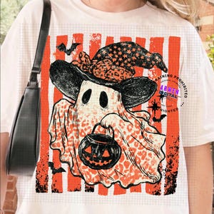 May include: A white t-shirt featuring a graphic of a ghost wearing a black witch hat with a leopard print pattern. The ghost holds a black jack-o'-lantern bucket. The background has orange vertical stripes and the text ARWEN DIGITAL.