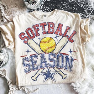 May include: Cream-colored t-shirt with a distressed graphic design. The design features the words "SOFTBALL SEASON" in red and blue, a yellow softball, crossed baseball bats, and a blue star.