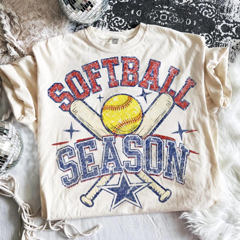 May include: Cream-colored t-shirt with a distressed graphic design. The design features the words "SOFTBALL SEASON" in red and blue, a yellow softball, crossed baseball bats, and a blue star.