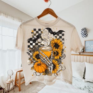 K&ouml;nnte beinhalten: Cremefarbenes T-Shirt mit einem Halloween-Motiv. Das Design zeigt eine dampfende Kaffeetasse, Sonnenblumen, einen K&uuml;rbis, Flederm&auml;use und eine Skeletthand. Das Shirt h&auml;ngt an einem Holzb&uuml;gel.