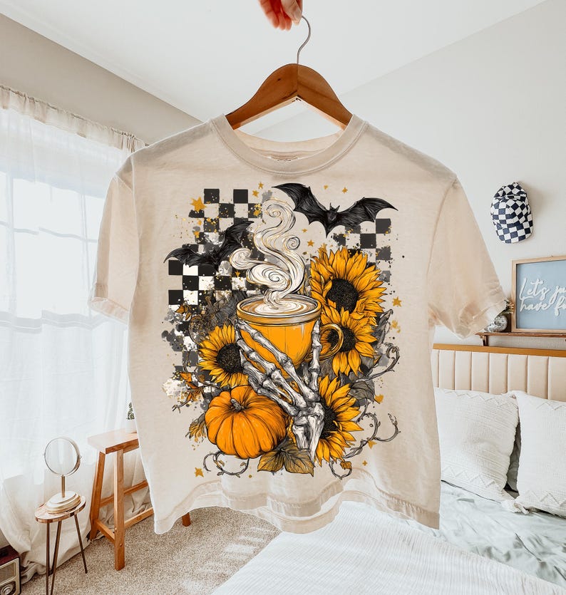 K&ouml;nnte beinhalten: Cremefarbenes T-Shirt mit einem Halloween-Motiv. Das Design zeigt eine dampfende Kaffeetasse, Sonnenblumen, einen K&uuml;rbis, Flederm&auml;use und eine Skeletthand. Das Shirt h&auml;ngt an einem Holzb&uuml;gel.