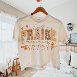 May include: A cream-coloured t-shirt featuring the words "I will PRAISE Him in every season" in a decorative font. The word "PRAISE" is designed with floral and plaid patterns. The shirt also includes the text "PSALM 146:2" and autumn-themed graphics.