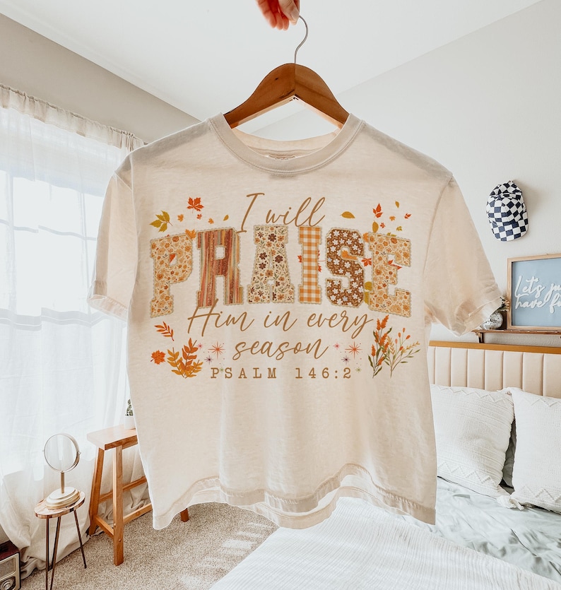 May include: A cream-coloured t-shirt featuring the words "I will PRAISE Him in every season" in a decorative font. The word "PRAISE" is designed with floral and plaid patterns. The shirt also includes the text "PSALM 146:2" and autumn-themed graphics.