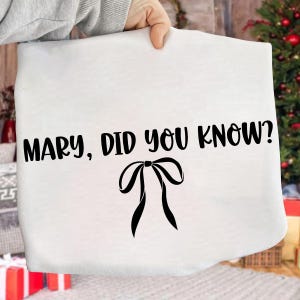 Może przedstawiać: Biały przedmiot z tkaniny z czarnym napisem &bdquo;MARY, DID YOU KNOW?&rdquo; i czarną grafiką kokardy. Przedmiot jest trzymany przez osobę, z choinką i prezentami w tle.
