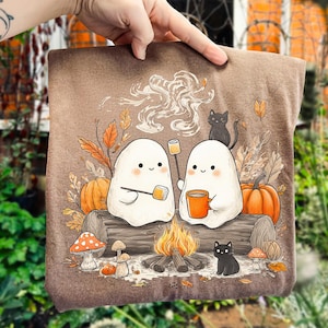 May include: A brown pillow with a whimsical illustration of two friendly ghosts roasting marshmallows over a campfire. The design includes pumpkins, black cats, mushrooms, and autumn leaves, creating a cozy fall theme.