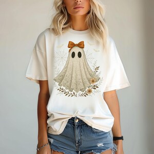 May include: Cream-colored t-shirt featuring a ghost design. The ghost is adorned with a bow and a lace-like pattern, surrounded by floral and leaf accents. The design includes spiderwebs and bats, perfect for Halloween.