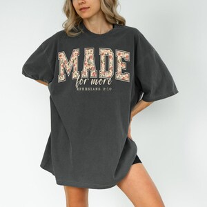 Op de afbeelding: Donkergrijs oversized T-shirt met de tekst "MADE for more" in een bloemenontwerp. De tekst "Ephesians 2:10" staat eronder. Het shirt heeft korte mouwen en een losse pasvorm.