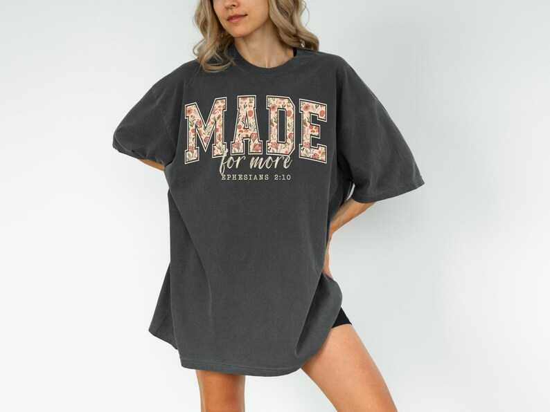 Op de afbeelding: Donkergrijs oversized T-shirt met de tekst "MADE for more" in een bloemenontwerp. De tekst "Ephesians 2:10" staat eronder. Het shirt heeft korte mouwen en een losse pasvorm.