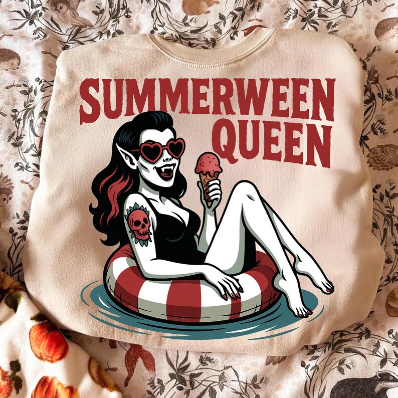 May include: Beige sweatshirt with the text "SUMMERWEEN QUEEN" in red. The graphic features a cartoon vampire with heart-shaped sunglasses, holding an ice cream cone while floating on a red and white striped inner tube. A skull tattoo is visible on the vampire's arm.