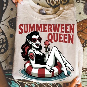 May include: Beige sweatshirt with the text "SUMMERWEEN QUEEN" in red. The graphic features a cartoon vampire woman in a black swimsuit, heart-shaped sunglasses, and a skull tattoo, floating in a red and white inner tube while holding an ice cream cone.
