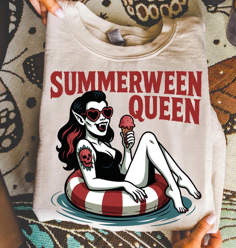 May include: Beige sweatshirt with the text "SUMMERWEEN QUEEN" in red. The graphic features a cartoon vampire woman in a black swimsuit, heart-shaped sunglasses, and a skull tattoo, floating in a red and white inner tube while holding an ice cream cone.
