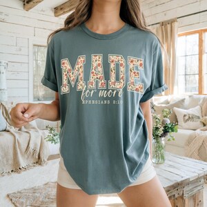 Op de afbeelding: Een tealkleurig t-shirt met de woorden "MADE" en "for more" in een bloemenpatroon. De tekst "Ephesians 2:10" is eronder gedrukt. Het shirt heeft opgerolde mouwen en een losse pasvorm.