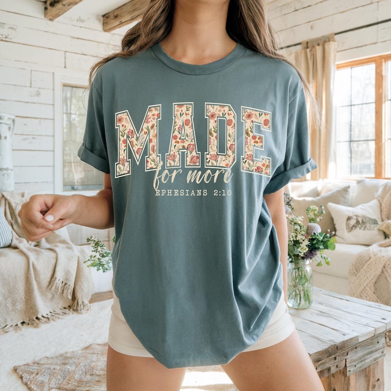Op de afbeelding: Een tealkleurig t-shirt met de woorden "MADE" en "for more" in een bloemenpatroon. De tekst "Ephesians 2:10" is eronder gedrukt. Het shirt heeft opgerolde mouwen en een losse pasvorm.