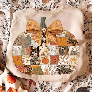 May include: Beige sweatshirt featuring a patchwork pumpkin design with a floral bow and a small skull. The pumpkin is composed of various floral and patterned squares, perfect for fall.
