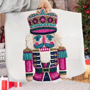 May include: A colorful, embroidered nutcracker design on a white background. The nutcracker features a crown, a pink and teal uniform, and a white beard. The design is held by a person.