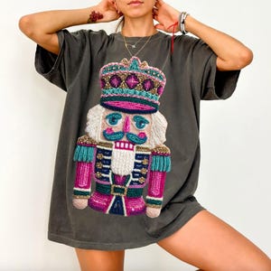 May include: Dark grey t-shirt featuring a colorful, embroidered nutcracker design. The nutcracker has a white beard, a pink and blue uniform, and a crown. The design is centered on the front of the shirt, creating a festive and eye-catching look.