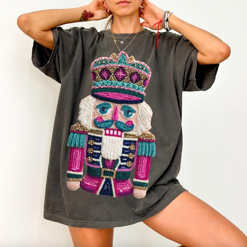 May include: Dark grey t-shirt featuring a colorful, embroidered nutcracker design. The nutcracker has a white beard, a pink and blue uniform, and a crown. The design is centered on the front of the shirt, creating a festive and eye-catching look.