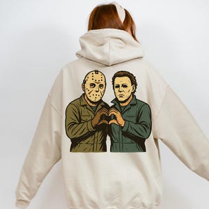 May include: Beige hoodie featuring a graphic of two horror movie characters, one wearing a hockey mask and the other with a pale face. They are making a heart shape with their hands. The hoodie has a drawstring hood.