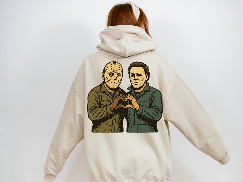 May include: Beige hoodie featuring a graphic of two horror movie characters, one wearing a hockey mask and the other with a pale face. They are making a heart shape with their hands. The hoodie has a drawstring hood.