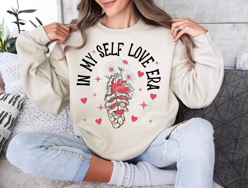 May include: Cream-colored sweatshirt with the text "IN MY SELF LOVE ERA" in black. The graphic features a skeletal hand holding a heart with flowers, surrounded by red hearts and stars. A cozy and stylish top.
