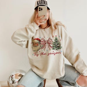 May include: Cream-colored sweatshirt featuring a Santa Claus graphic, a red and white striped bow, and a Christmas tree. The text "Tis the season" is printed below the graphics. The person in the image is wearing a white cap with the word "KARMA" on it.