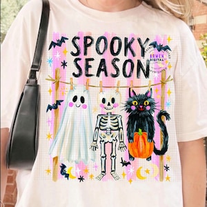 K&ouml;nnte beinhalten: Ein cremefarbenes T-Shirt mit einer Grafik von einem Geist, einem Skelett und einer schwarzen Katze mit K&uuml;rbis, die an einer W&auml;scheleine h&auml;ngen. Der Text SPOOKY SEASON ist von rosa Streifen, gelben Sternen und schwarzen Flederm&auml;usen umgeben.