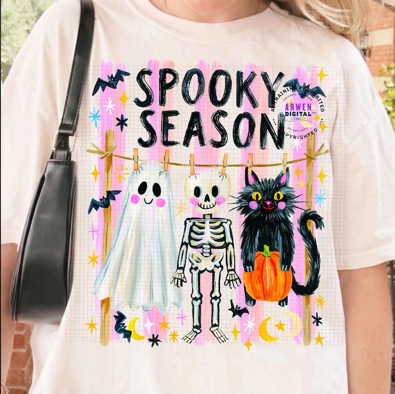 K&ouml;nnte beinhalten: Ein cremefarbenes T-Shirt mit einer Grafik von einem Geist, einem Skelett und einer schwarzen Katze mit K&uuml;rbis, die an einer W&auml;scheleine h&auml;ngen. Der Text SPOOKY SEASON ist von rosa Streifen, gelben Sternen und schwarzen Flederm&auml;usen umgeben.