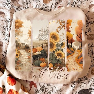 May include: Beige sweatshirt with a fall-themed graphic. The design features four vertical panels with images of sunflowers, pumpkins, and a pathway. The words "Fall Vibes" are written in a cursive font across the bottom of the graphic.